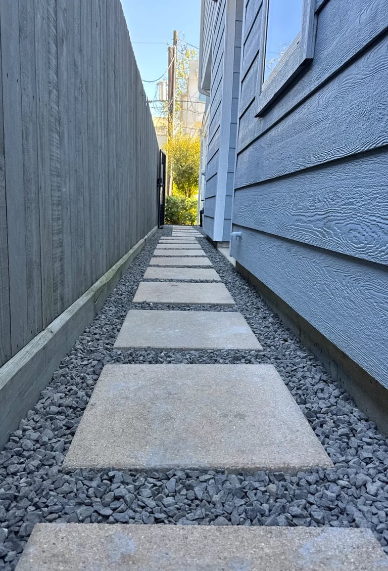 Narrow walkway between two modern blue-grey buildings with concrete stepping stones and gravel, leading toward a distant church spire and trees
