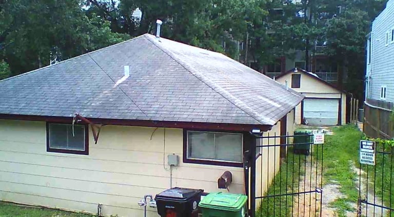 Small single-story house with gray roof, cream-colored siding, and dark trim surrounded by trees and a metal fence.