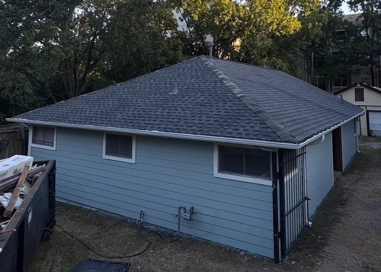 Light blue single-story house with dark gray shingle roof and front porch, surrounded by trees