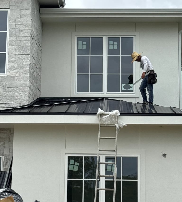 Worker in safety gear standing on roof of two-story house with metal roofing and large windows, using a ladder.