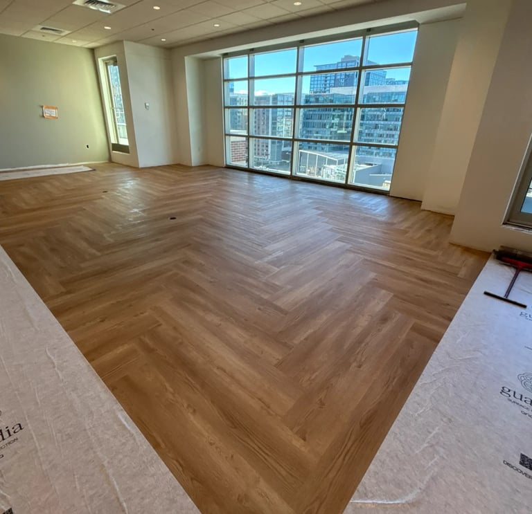 Empty modern office space with herringbone wood flooring, large windows overlooking city skyline, and white walls with recessed lighting