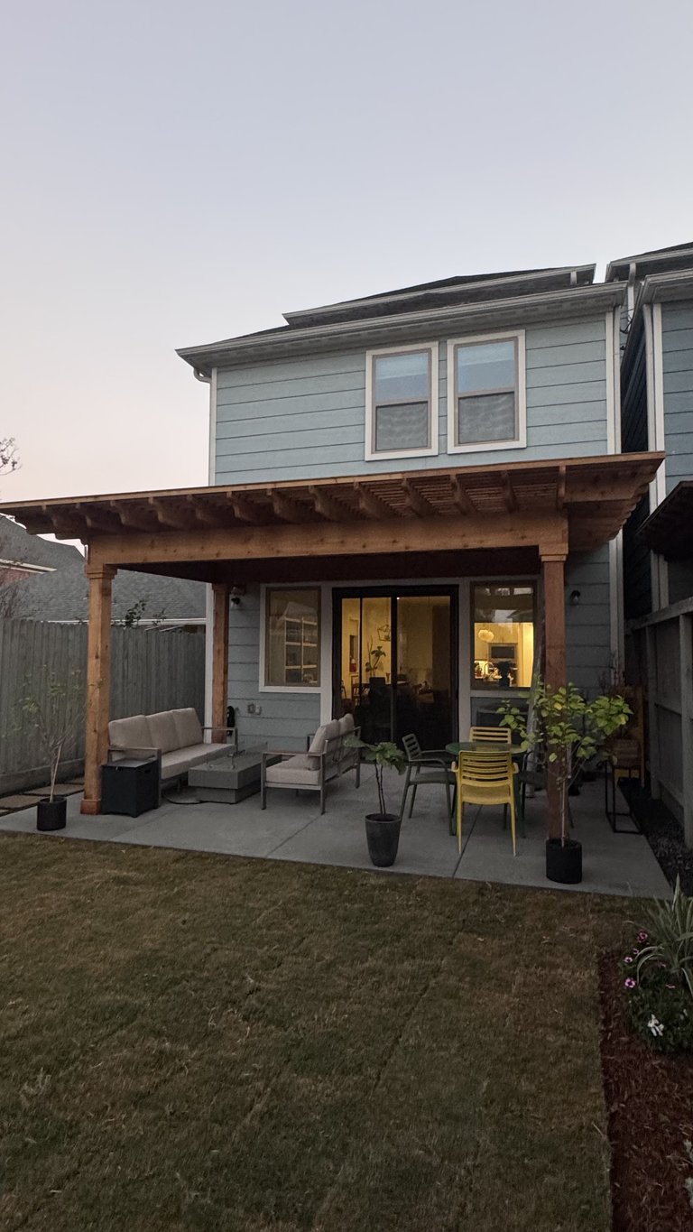 Modern two-story light blue house with wooden pergola patio, outdoor seating, and landscaping at dusk
