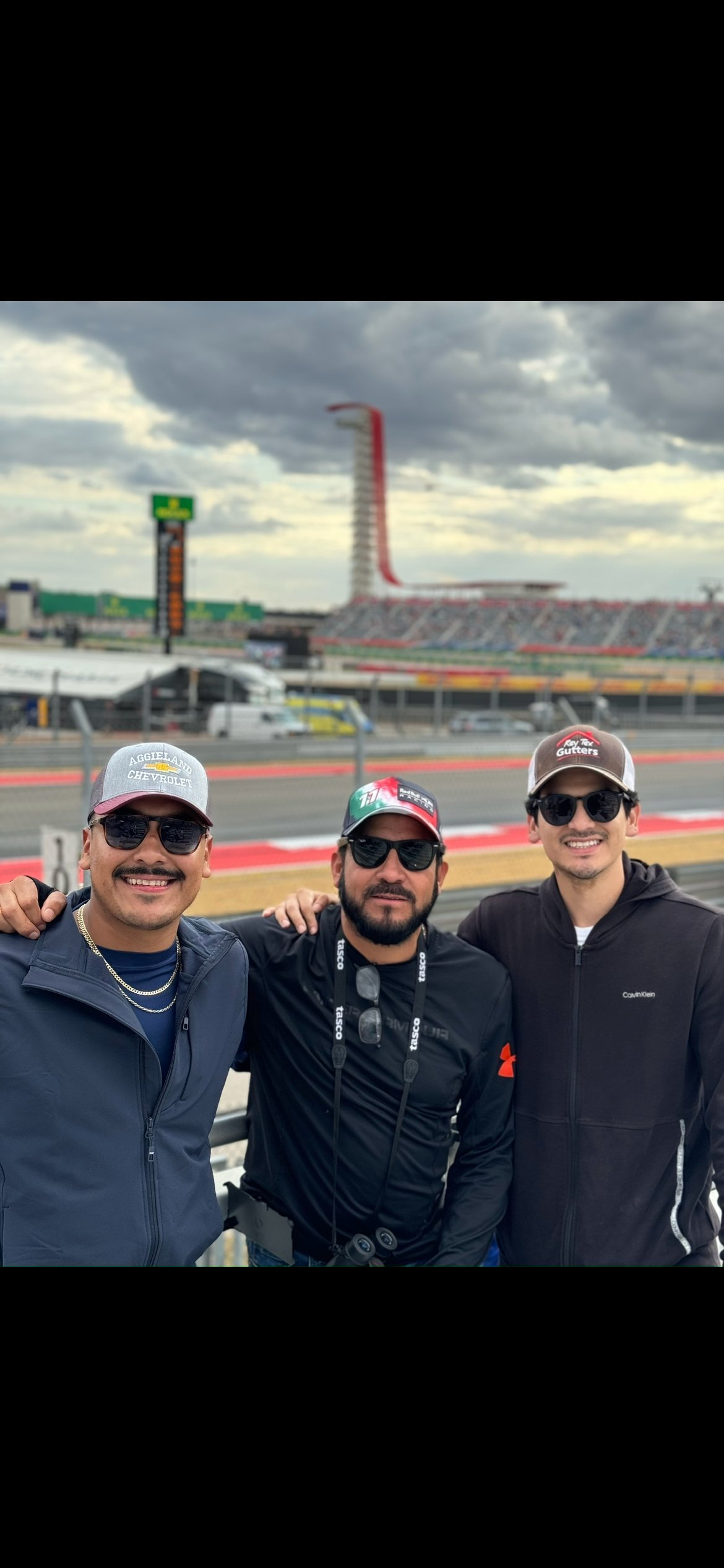 Three men wearing sunglasses and baseball caps smile at a racing track with grandstands and a tall red structure in the background.