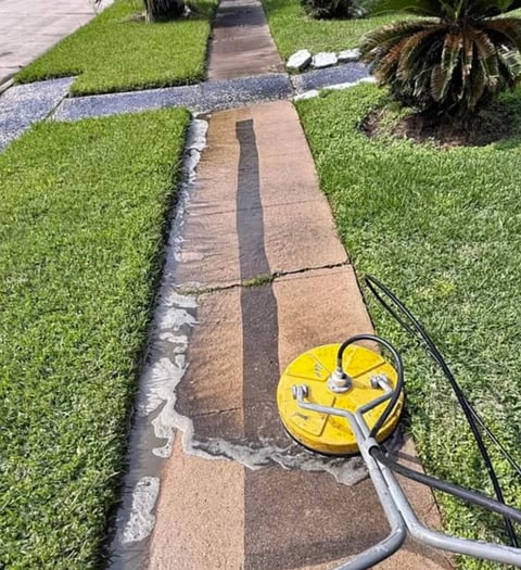 A yellow pressure washer with hose cleaning a stone pathway between manicured green lawns in a residential yard