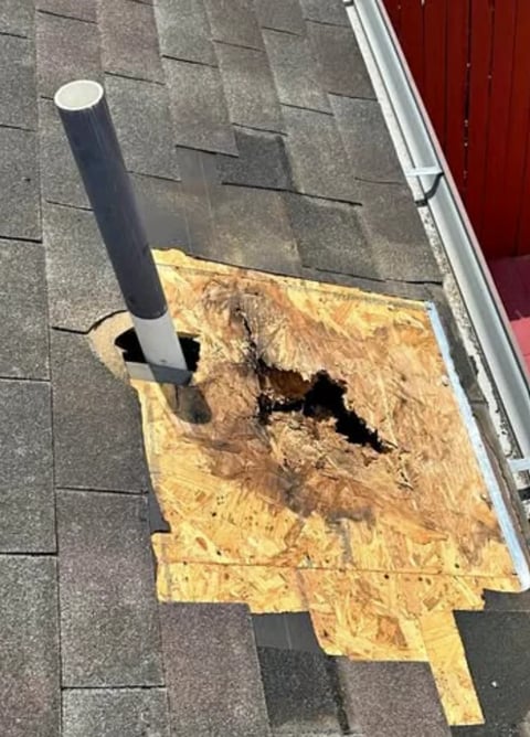 Roof damage with plywood patch over hole next to metal vent pipe and red gutter trim.