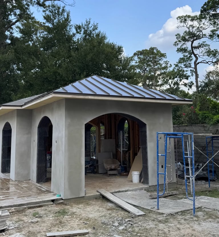 Modern white concrete house under construction with metal roof, arched doorway, and blue scaffolding, surrounded by trees