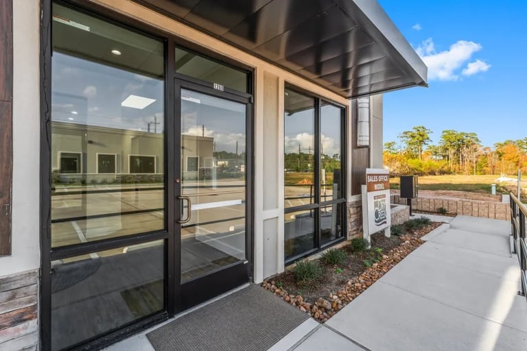 Modern storefront with black-framed glass doors and windows on a concrete patio overlooking a wooded area under clear blue sky