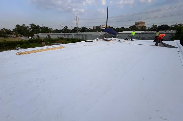 Workers installing white roofing membrane on a large flat commercial roof with trees and transmission tower in background