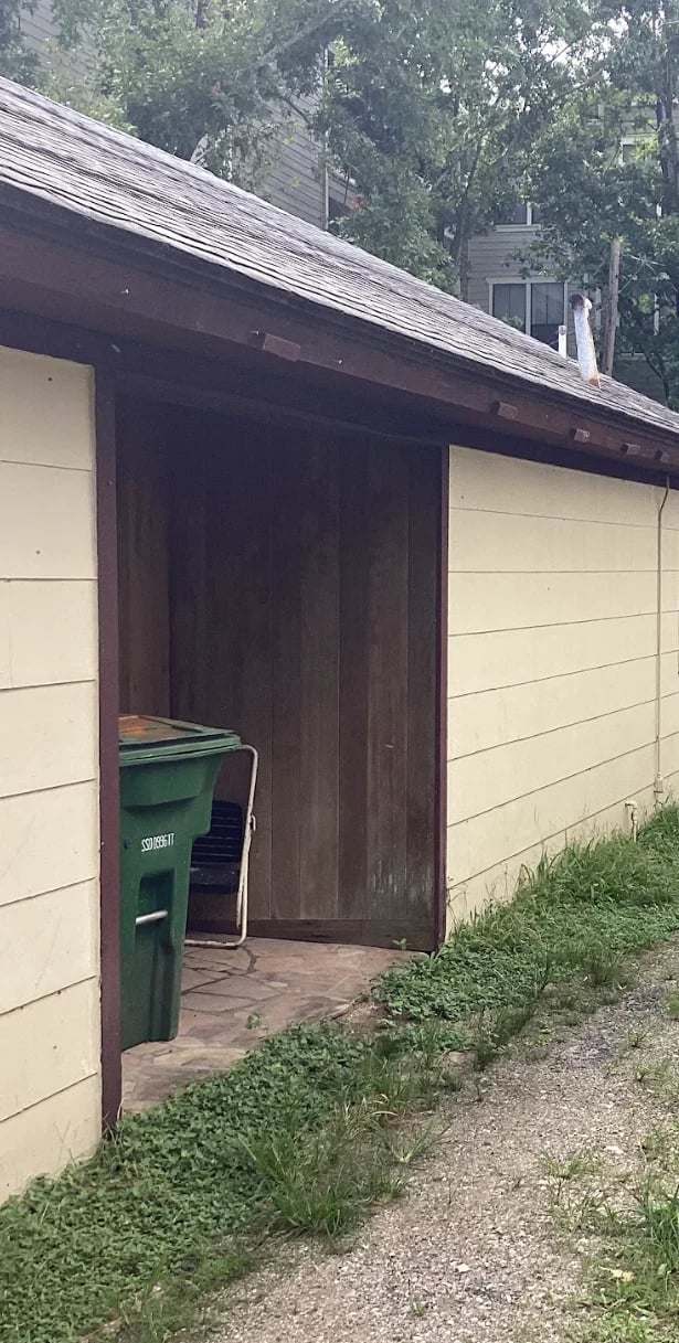 A wooden garage with cream and dark brown siding, open double doors, and a green trash bin outside, surrounded by overgrown grass
