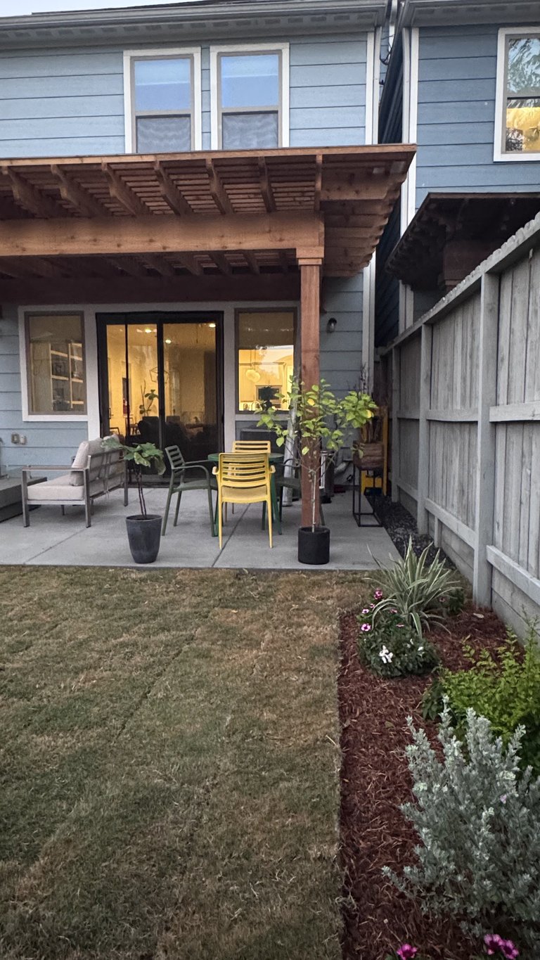 Modern blue house with wooden pergola over patio area, featuring seating, potted plants, and yard landscaping
