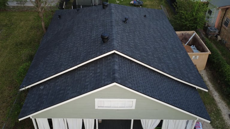 Aerial view of a residential house with dark gray shingle roof and garage, surrounded by green lawn and trees