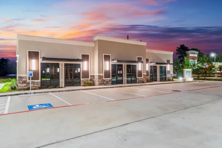 Modern commercial building with large storefronts at dusk, featuring tan exterior, glass doors, and landscaping under a vibrant sunset sky