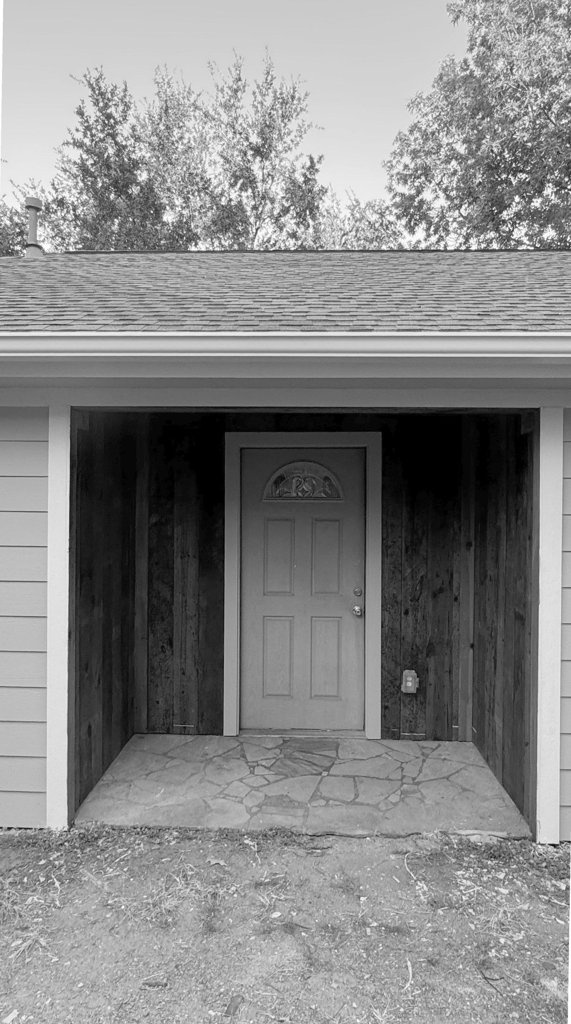 Black and white photo of a house entrance with a gray door under a covered porch, surrounded by weathered wood siding and trees in the background