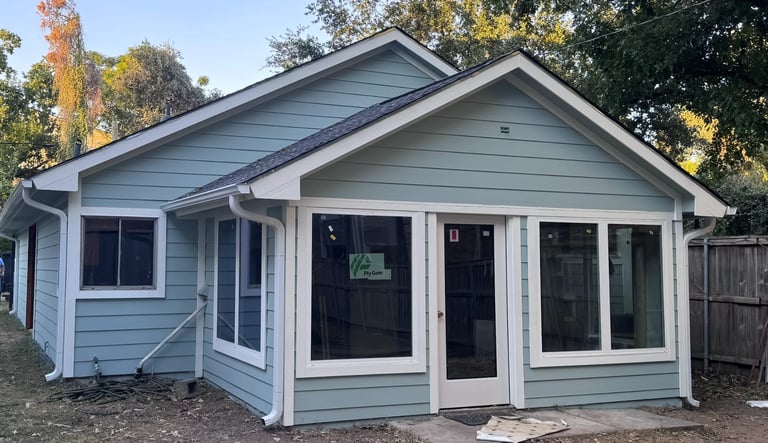 Light blue single-story house with white trim, large front windows, covered porch, and leafy trees in the background during fall.