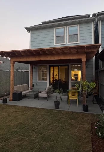 Modern two-story house with blue-gray siding, wooden pergola patio, glass doors, outdoor furniture, and landscaping at dusk