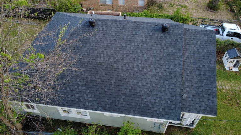 Aerial view of a residential house with a dark gray asphalt shingle roof and white gutters, surrounded by trees and landscaping.