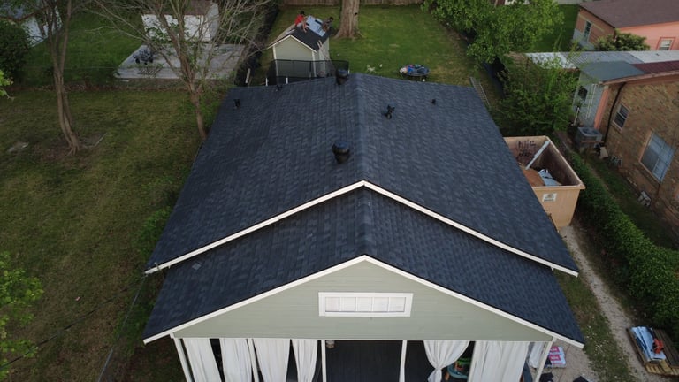 Aerial view of a house with dark gray shingle roof and white trim, surrounded by green lawn and trees