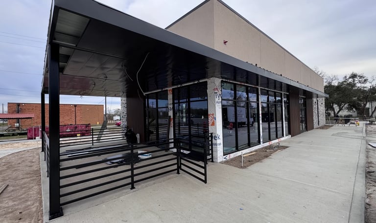 Modern commercial storefront with black metal awning, glass doors and windows, concrete sidewalk, and adjacent industrial buildings under cloudy sky