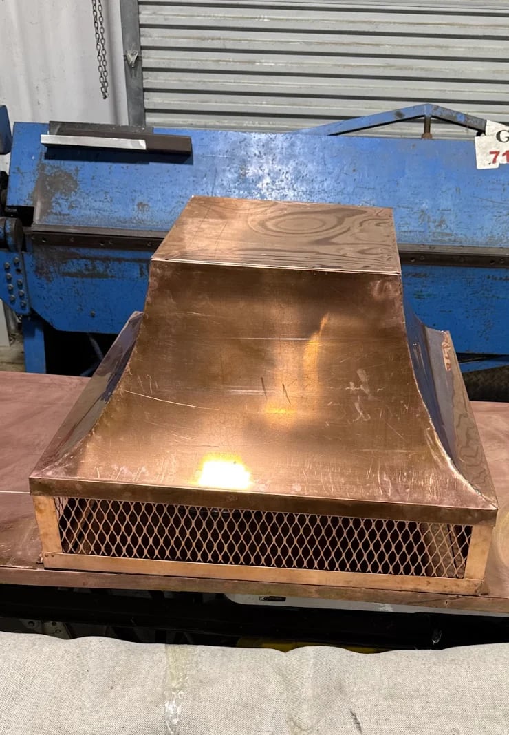Copper kitchen range hood with curved design and mesh vent sitting on work surface with blue equipment in background