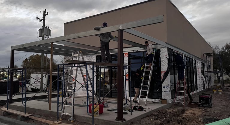 Construction workers on ladders building a metal frame structure next to a brown industrial building under cloudy skies