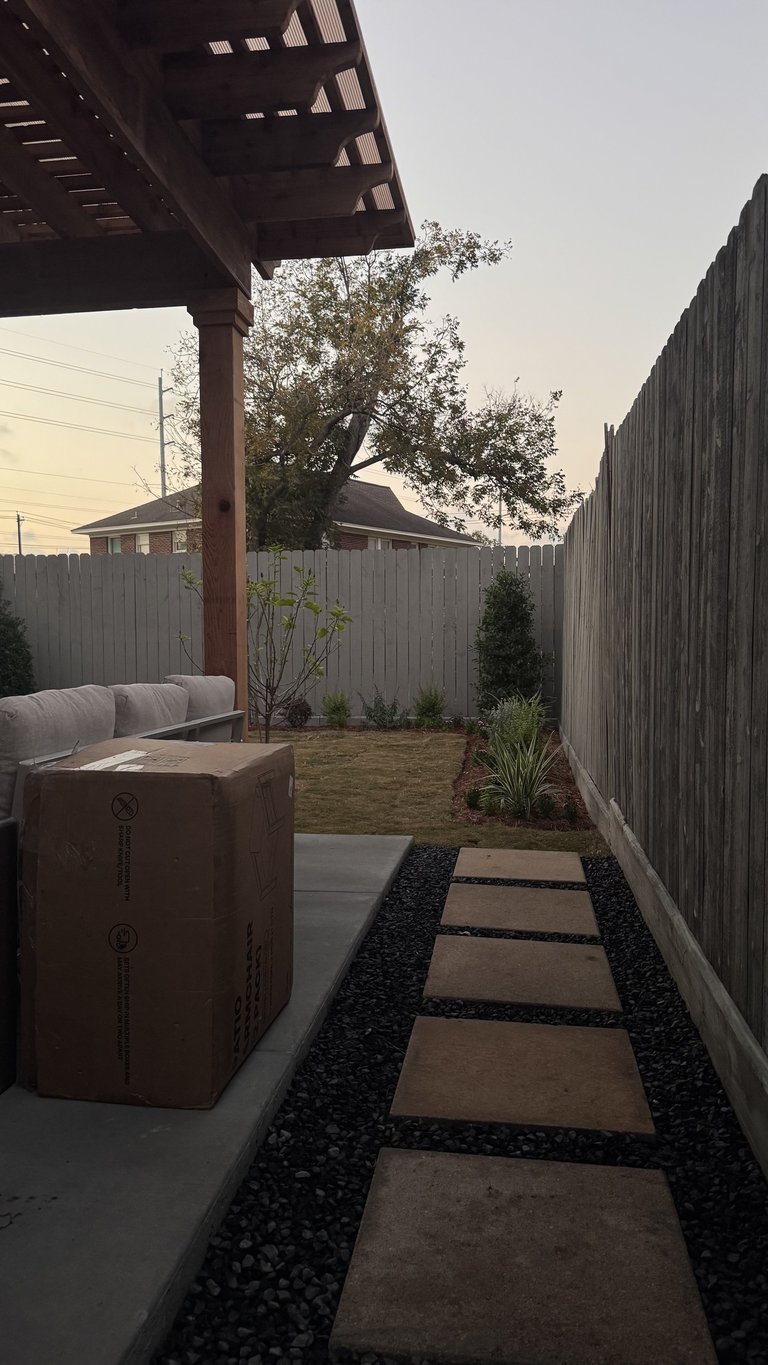 Narrow backyard with stepping stone pathway, gray metal fences, pergola overhead, and planted landscape areas at dusk