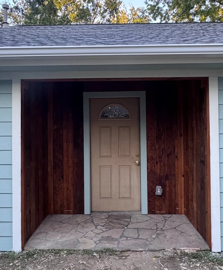 Front entrance of a house with a tan wooden door, dark wood siding, stone patio, and white trim under a gray roof with trees in the background