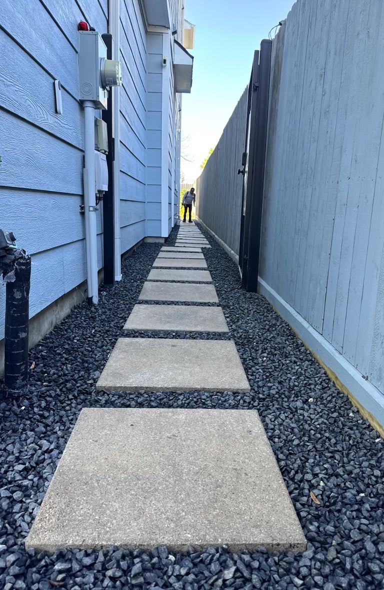 Narrow walkway between two buildings with stepping stones and gravel, blue house on left, gray fence on right