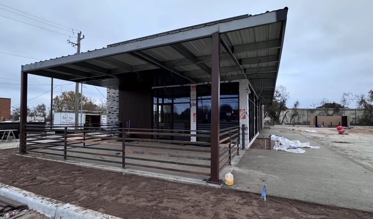 Modern single-story building with steel frame, metal roof, and large windows on a construction site with dirt ground and scattered patches of snow